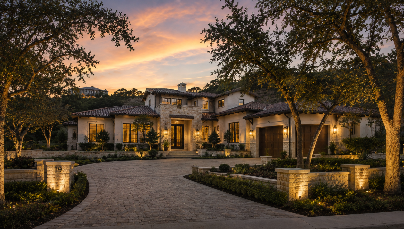 A stone and stucco residence framed by mature trees at sunset, with warm light in its windows.